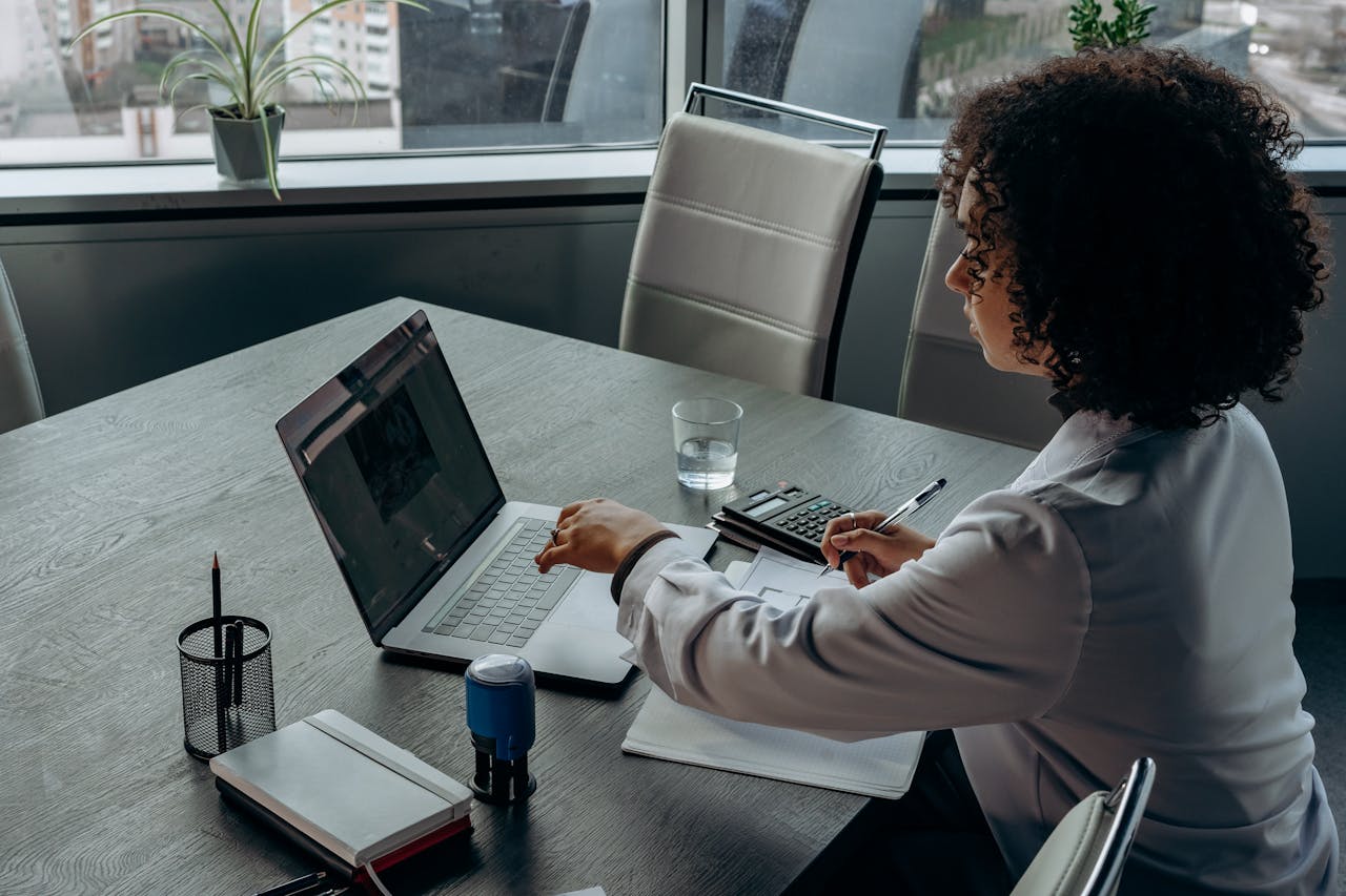 A businesswoman with curly hair works on a laptop in a modern office environment, focusing on tasks.